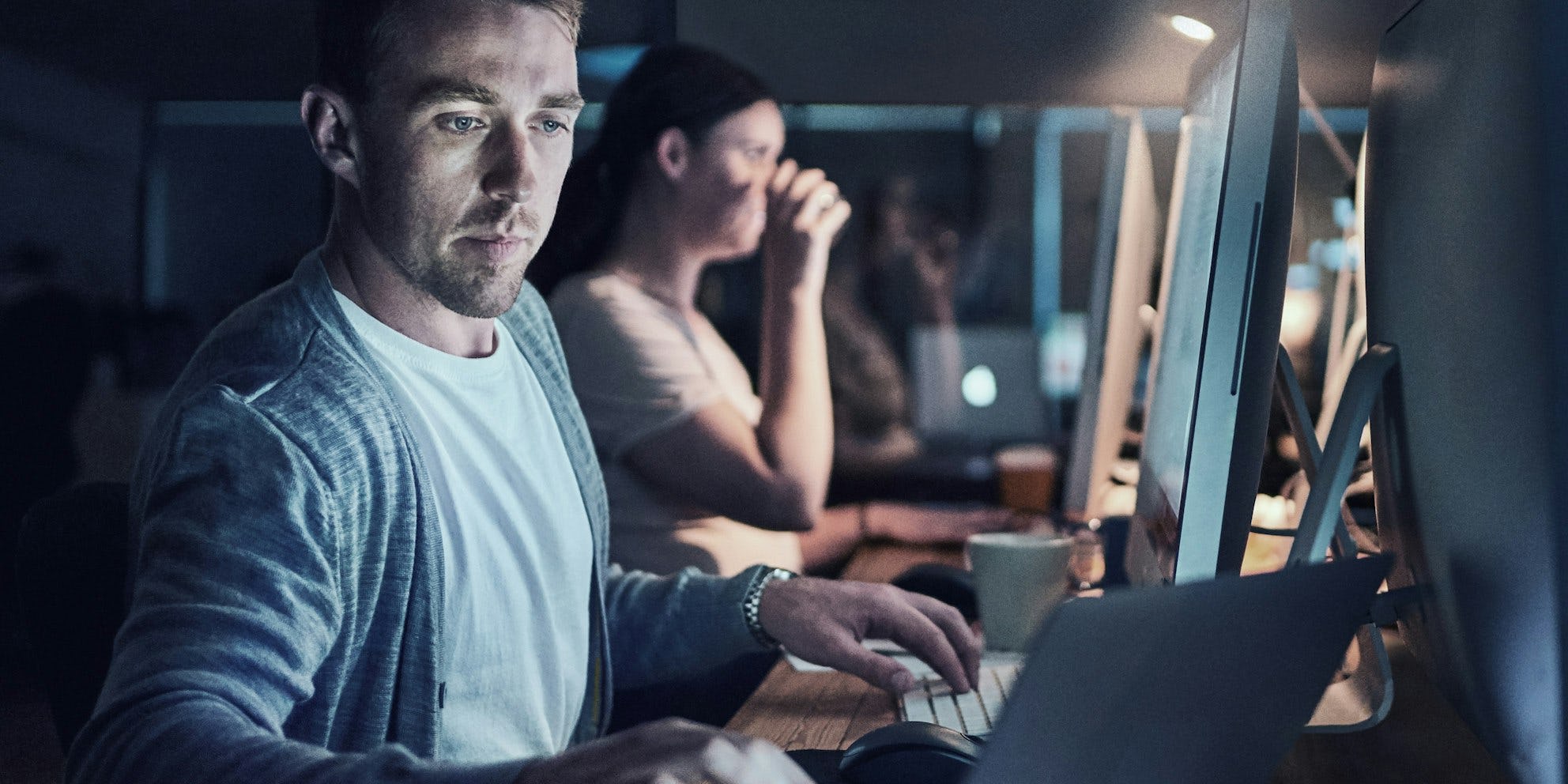 Een man en vrouw werken achter hun computer op kantoor. De man kijkt naar zijn laptop en de vrouw drinkt koffie terwijl ze naar haar computer scherm kijkt.