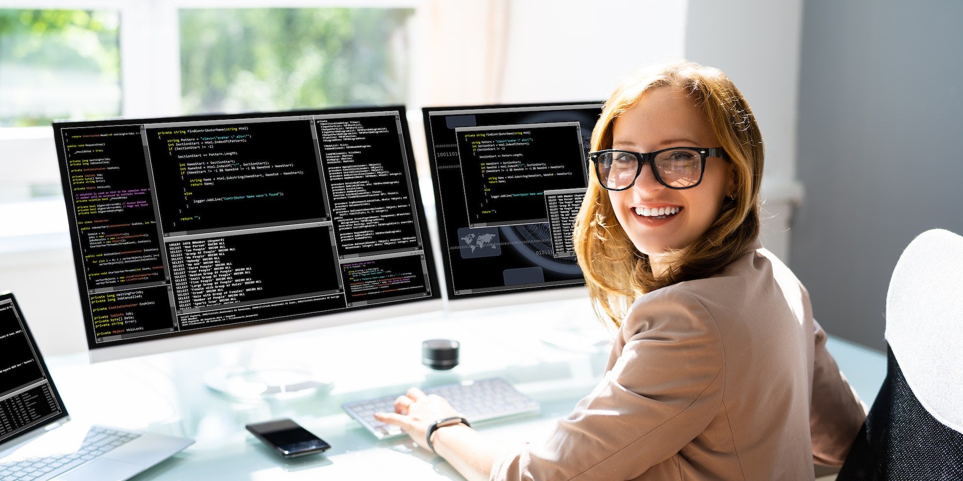 Een vrouw kijkt vrolijk de camera in met achter haar twee computerschermen met codeertaal erop.